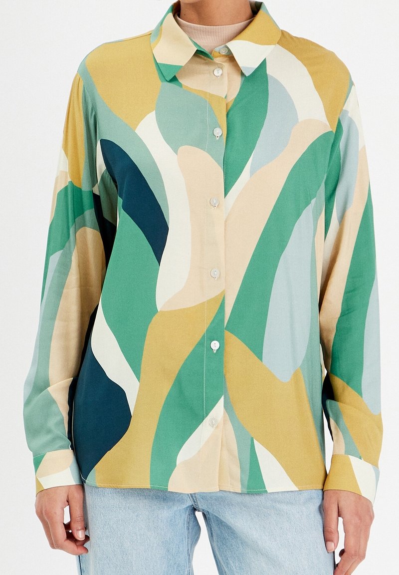 Chemise manches longues à boutons avec des motifs abstraits en vagues de couleur verte, jaune, beige et bleue, portée avec un jean bleu clair.