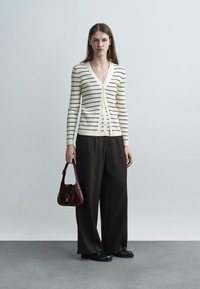 Moteris dėvi kreminės ir juodos spalvos dryžuotą kardiganą, plačias juodas kelnes, juodus batus, laiko mažą tamsiai raudoną rankinę, stovi ant pilko grindinio.