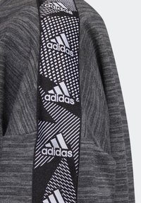 Sudadera gris con un panel lateral negro que presenta el logo blanco de Adidas y patrones geométricos. La tela parece suave con un acabado texturizado.