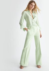Blusa di raso verde chiaro con maniche lunghe e colletto, abbinata a pantaloni a vita alta coordinati con un accento dorato sulla cintura.