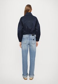 Marineblå, kort jakke med innsnørt midje, høy krage og puffermer, sammen med lyseblå rettskårne jeans med opprettede mansjetter.