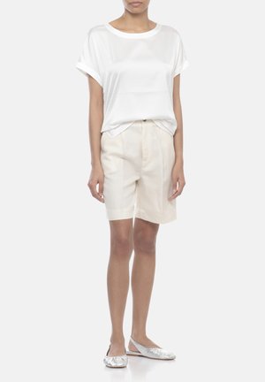 Donna con top bianco a maniche corte largo, pantaloncini beige su misura fino al ginocchio e scarpe slingback piatte color argento metallizzato che stanno su uno sfondo bianco.