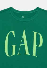 Zelené bavlněné tričko s krátkým rukávem, s velkým světle zeleným logem "GAP" potištěným vpředu a klasickým kulatým výstřihem.