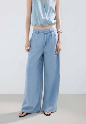 Mujer con pantalones vaqueros anchos azul claro, camiseta sin mangas azul claro drapeada, sandalias negras, pulsera y anillo plateados, de pie sobre un fondo liso.