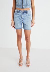 Gaišā zila džinsa šorti ar saplēstu apakšmalu, kombinēti ar īsu džinsa vestu. Modelis valkā baltas sandales ar siksniņām.