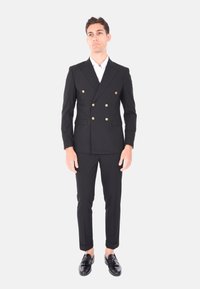 Giacca nera doppiopetto con bottoni dorati, pantaloni abbinati, vestibilità sartoriale e una camicia bianca sotto. Scarpe nere lucide.