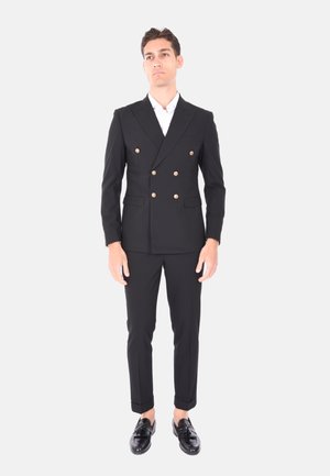 Giacca nera doppiopetto con bottoni dorati, pantaloni abbinati, vestibilità sartoriale e una camicia bianca sotto. Scarpe nere lucide.