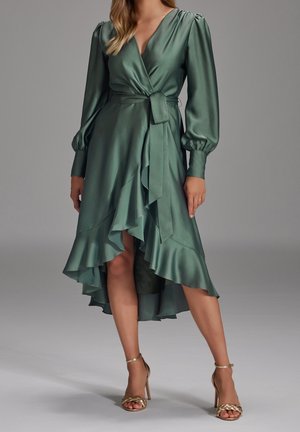 Femme portant une robe portefeuille en satin vert sauge avec des manches longues bouffantes et un ourlet à volants, associée à des sandales à talons hauts dorées tressées.