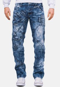 Jeans in denim blu con un lavaggio sbiadito, cuciture diagonali e verticali, molteplici tasche e dettagli in metallo decorativi.