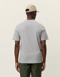 Sivý bavlnený tričko s krátkymi rukávmi a okrúhlym výstrihom, nosené so zelenými pruhovanými nohavicami, zobrazené zozadu, bez viditeľných log.