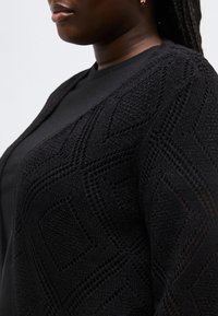 Nahaufnahme einer Person, die eine schwarze, strukturierte Strickjacke über einem schwarzen Shirt trägt, mit Fokus auf das geometrische Muster und die Details des Ohrrings.