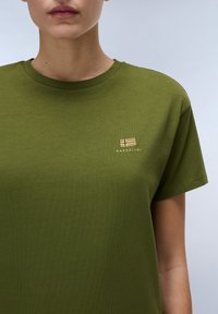 Olivengrünes T-Shirt aus weichem Stoff mit Rundhalsausschnitt und kurzen Ärmeln. Es besitzt ein dezentes, goldfarbig gesticktes Logo auf der linken Brust.