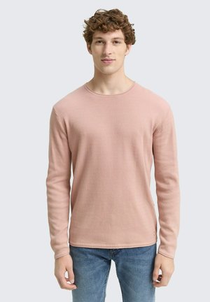 Langsleeve trui met ronde hals in een lichtroze, gestructureerde brei-stof. Een subtiele logo-accent op de linksonder. Gecombineerd met blauwe spijkerbroek.