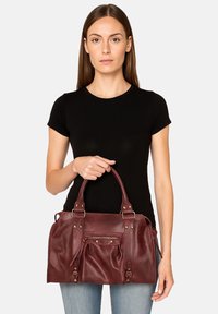 Borsa in pelle bordeaux con manici arrotondati, tasca frontale con zip e dettagli in ottone. Texture liscia e design strutturato.