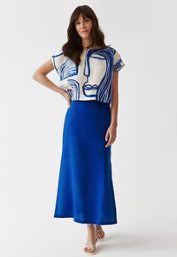 Lněný crop top s modrým abstraktním liniovým uměním, zkombinovaný s dlouhou, jednobarevnou modrou sukní. Modelka má na sobě bílé sandály na popruh.