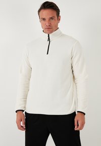 Hvid fleece-pullover med høj krave og sort lynlås. Har lange ærmer med sorte manchetter og en glat tekstur.
