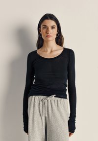 Žena s dugom tamnom kosom nosi crnu majicu dugih rukava i svijetlosive hlače s vrpcama, stojeći uz jednobojnu svijetlu pozadinu.