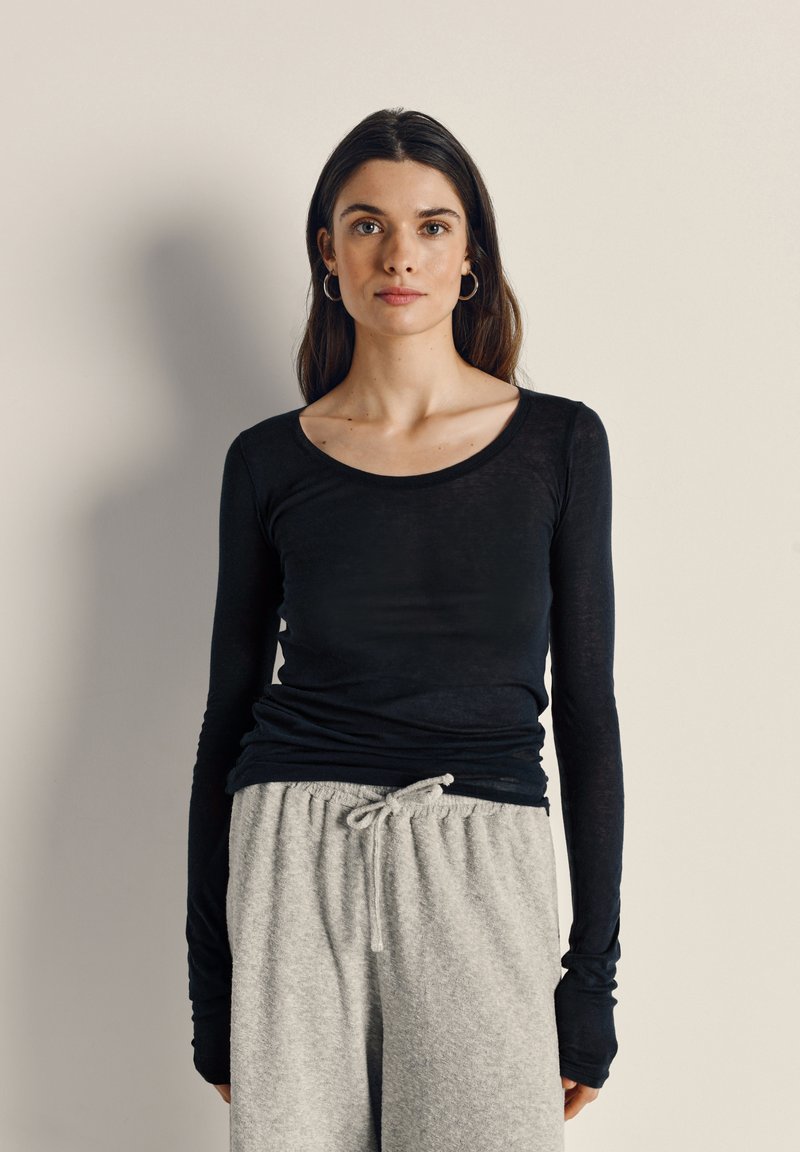 Žena s dugom tamnom kosom nosi crnu majicu dugih rukava i svijetlosive hlače s vrpcama, stojeći uz jednobojnu svijetlu pozadinu.