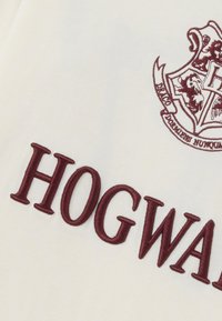 Witte stof met bordeauxrood geborduurde tekst "HOGWARTS" en een gedeeltelijk Hogwarts-wapen met vier dieren en een banner.