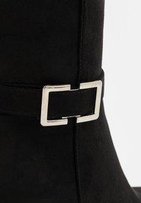 Détail d'une botte en daim noir avec une boucle rectangulaire en argent et une sangle cousue autour de la cheville sur un fond blanc uni.