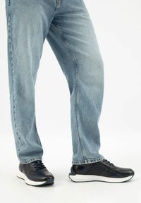 Muž v světle modrých džínách a černých teniskách s bílými podrážkami, stojící před jednoduchým bílým pozadím.