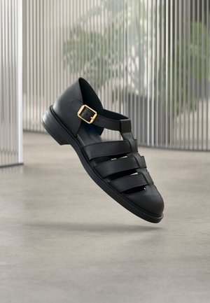 Melna ādas sandaļa ar noslēgtu pirkstu, trim horizontālām siksniņām un zelta sprādzi potītes siksniņā, peld virs betona grīdas telpās.