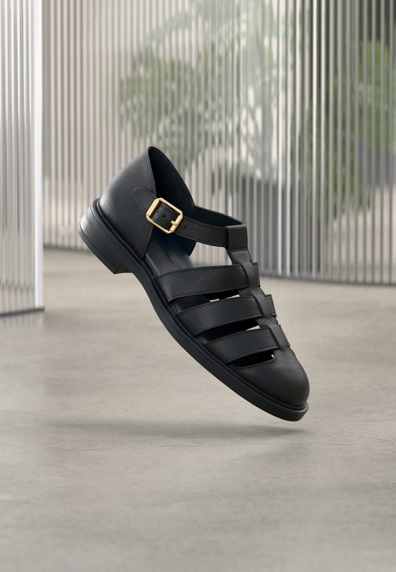 Μαύρο δερμάτινο σανδάλι με κλειστή μύτη, τρεις οριζόντιες λουρίδες και χρυσή αγκράφα στον ιμάντα του αστραγάλου, αιωρείται πάνω από το τσιμεντένιο πάτωμα σε εσωτερικό χώρο.