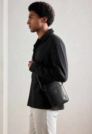 Mann in dunkler Jacke und hellen Hosen, der eine schwarze Lederumhängetasche trägt und im Profil vor einem einfarbigen Hintergrund steht.