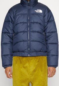 Mörkblå pufferjacka med kviltad design, hög krage, framdragkedja och logotypdetalj. Har elastiska ärmslut och nederkant för en tät passform.