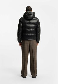 Sort polstret jakke med hætte, der har en glansfuld overflade og elastiske ærmer, båret over ternede bukser. Enkel, funktionel design.