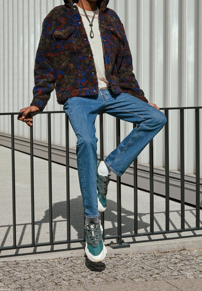 Personne assise sur une rambarde métallique noire portant un jean bleu, des baskets vertes et blanches, une veste polaire à motifs et une chemise blanche.
