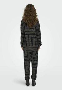 Zwarte outfit met een geometrisch patroon van witte lijnen. De top heeft lange mouwen en de broek heeft een losse fit. Zwarte blokhakken zijn zichtbaar.