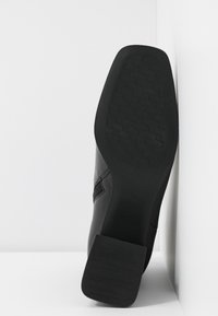 Semelle noire d'une bottine à talon avec une semelle texturée, positionnée verticalement contre un coin de mur et de sol blanc.