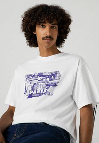 Valkoinen puuvillat-paita, jossa on violetti grafiikkaprintti Pariisin maamerkeistä ja tekstistä. Lyhyet hihat, pyöreä kaula-aukko ja rento malli.