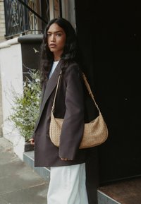 Beige geweven schoudertas met een gebogen vorm, aangevuld met een lange schouderriem, gecombineerd met een donkere oversized blazer en lichte, wijde broek.
