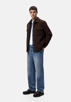 Jonge man staat met handen in de zakken, draagt een bruine overshirt, donkerblauw overhemd, wit T-shirt, lichtblauwe spijkerbroek en zwarte schoenen.