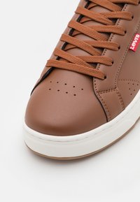 Barna bőr sneaker perforált orral, világosbarna fűzőkkel és fehér gumi talppal. Piros logó részletek a fűzők mellett. Az egész sima textúrájú.