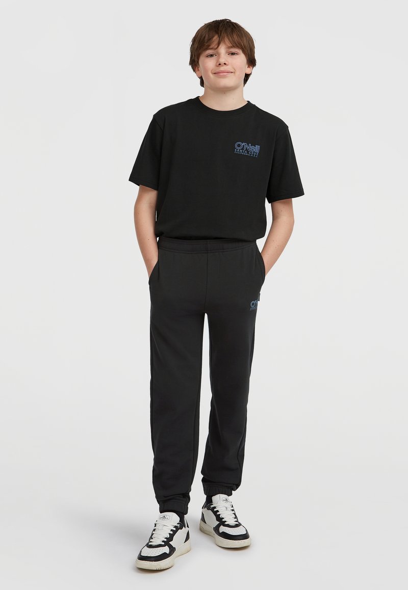 Zwarte katoenen t-shirt met blauw logo, gecombineerd met zwarte jogginbroeken met een elastische tailleband en zijzakken, afgemaakt met zwart-witte sneakers.