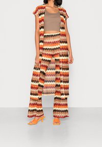 Conjunto de punto con un diseño de zigzag multicolor, que incluye un cárdigan largo abierto, una camiseta corta y pantalones acampanados en tonos tierra. Sandalias de tacón naranjas.