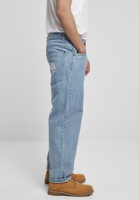 Lyseblå denimjeans med en avslappet passform, med baklommer og en hvit brodert logo på høyre lomme. Kombinert med brune støvler.