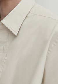 Beżowa, bawełniana koszula z ostrym kołnierzykiem, charakteryzująca się gładką fakturą i precyzyjnym wykończeniem szwów wzdłuż zapięcia z przodu.