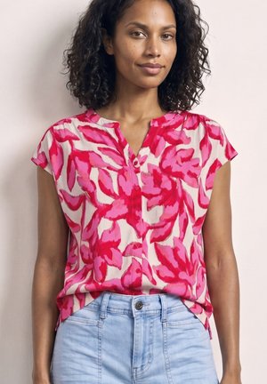 Vrouw draagt een kortarmige button-up blouse met een roze en rode abstracte bloemenprint, ingestopt in lichtblauwe spijkerbroek.