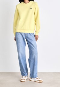 Felpa gialla in pile con polsini e orlo a costine, abbinata a jeans larghi blu chiaro e sneakers bianche con accenti marroni.