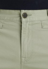 Záber na pás svetlozelených nohavíc s kovovým gombíkom s vyžehleným nápisom "PME Legend Jeans" a časťou čierneho rebrovaného trička vyššie.