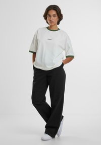 Camiseta blanca con cuello acanalado verde, corte casual, combinada con pantalones negros y zapatillas blancas. Presenta un logo bordado en el pecho.