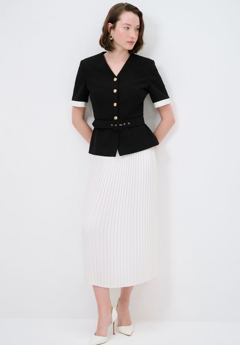 Schwarzer taillierter Blazer mit goldenen Knöpfen und einem taillierten Gürtel, kombiniert mit einem fließenden weißen Faltenrock. Weiße Absätze vervollständigen das Outfit.