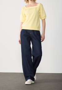 Person som bär en kortärmad topp i gult med ögla-detaljer, mörka vida jeans och vita sneakers, stående på ett grått golv.