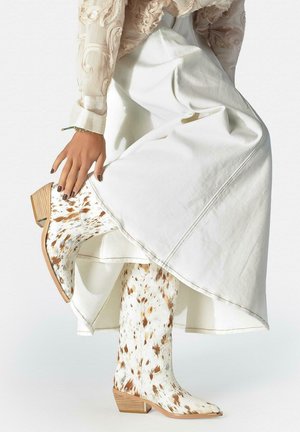 Persoon in witte rok heft been op, toont witte en bruine gevlekte cowboylaarzen met houten hakken en een doorschijnende geborduurde blouse mouw.