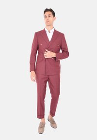 Abito bordeaux doppiopetto in tessuto testurizzato, con rever a punte e bottoni dorati. Pantaloni slim-fit con orli sovrapposti e calzature marrone chiaro.