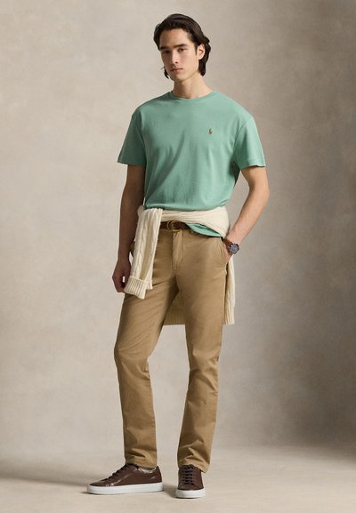 Νέος άνδρας φοράει γαλαζοπράσινο μπλουζάκι, μπεζ παντελόνι, καφέ αθλητικά παπούτσια, μπεζ πουλόβερ δεμένο γύρω από τη μέση και ρολόι χειρός στον αριστερό καρπό.